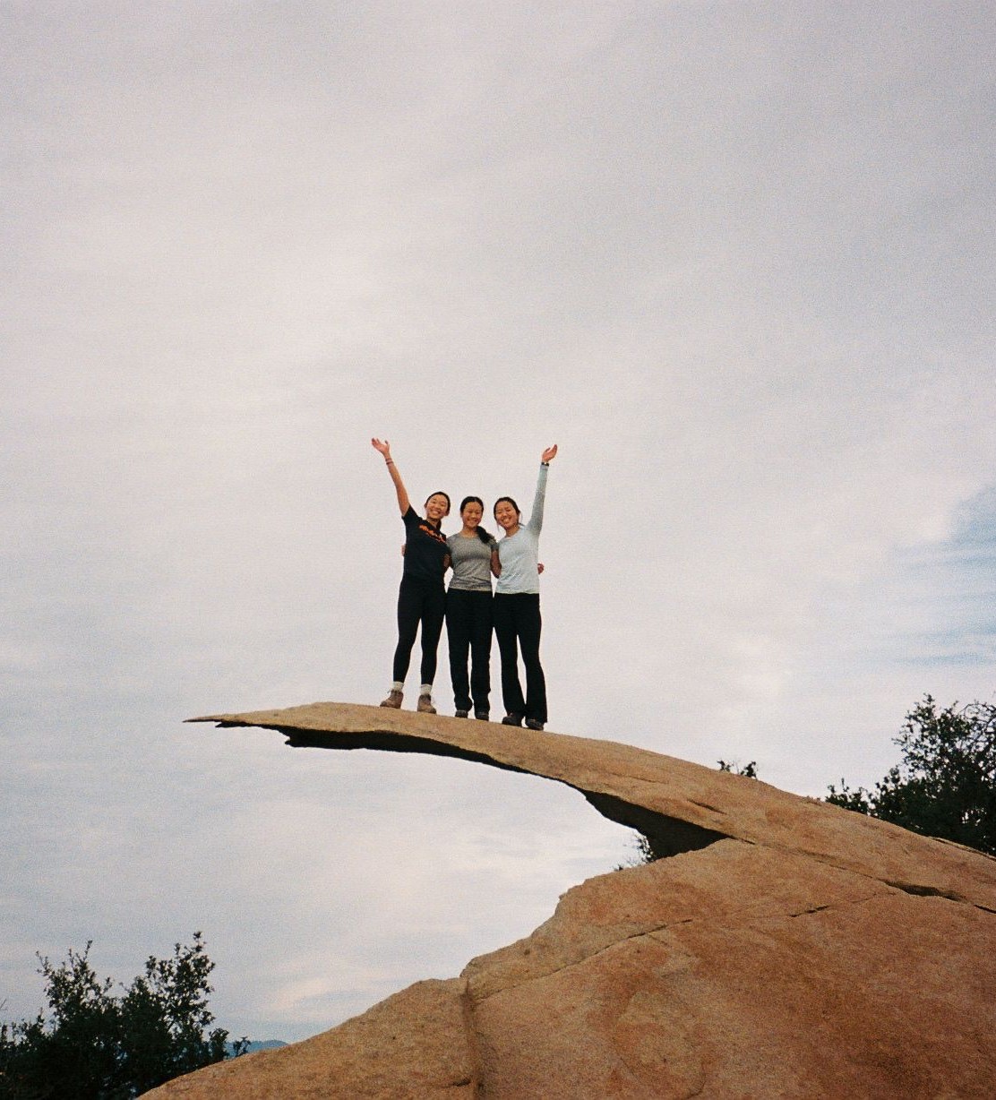 three girls hugging and standing on a thin slab of rock jutting out from a mountain